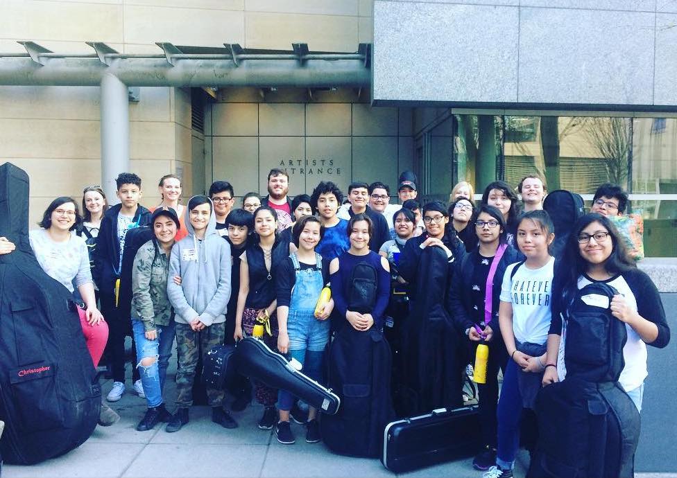 Smiling student musicians gathered in front of a concert hall with their instruments.
