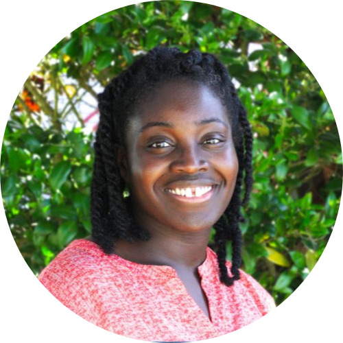 Lianna Olds, a Black woman with black hair and brown eyes and a big smile stands in front of greenery.
