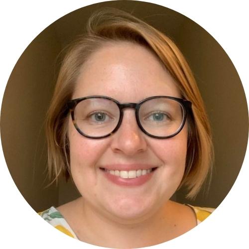 Tiffany Brace, a white woman with light colored hair, is smiling and wearing glasses. The background behind her is dark.